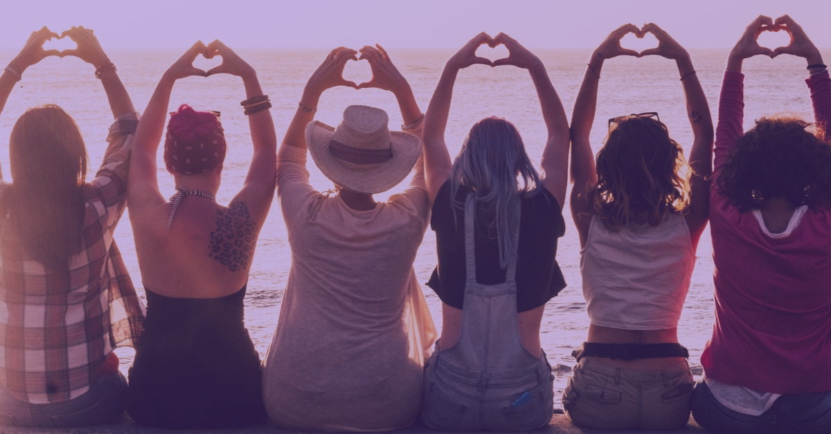 the backs of several women sitting in a line on the beach with their arms outstretched over their heads making hearts with their hands