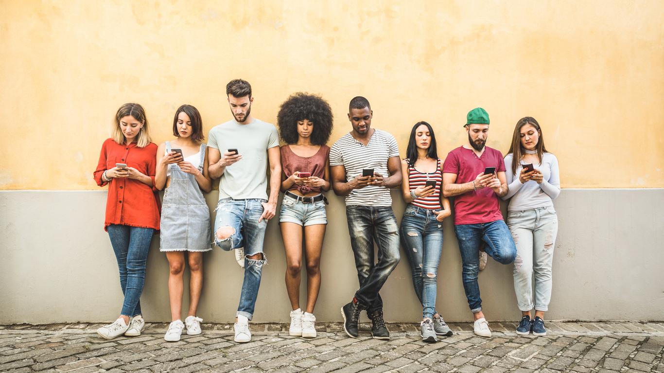 Photo shows a group of young people all standing by a wall looking at their phones