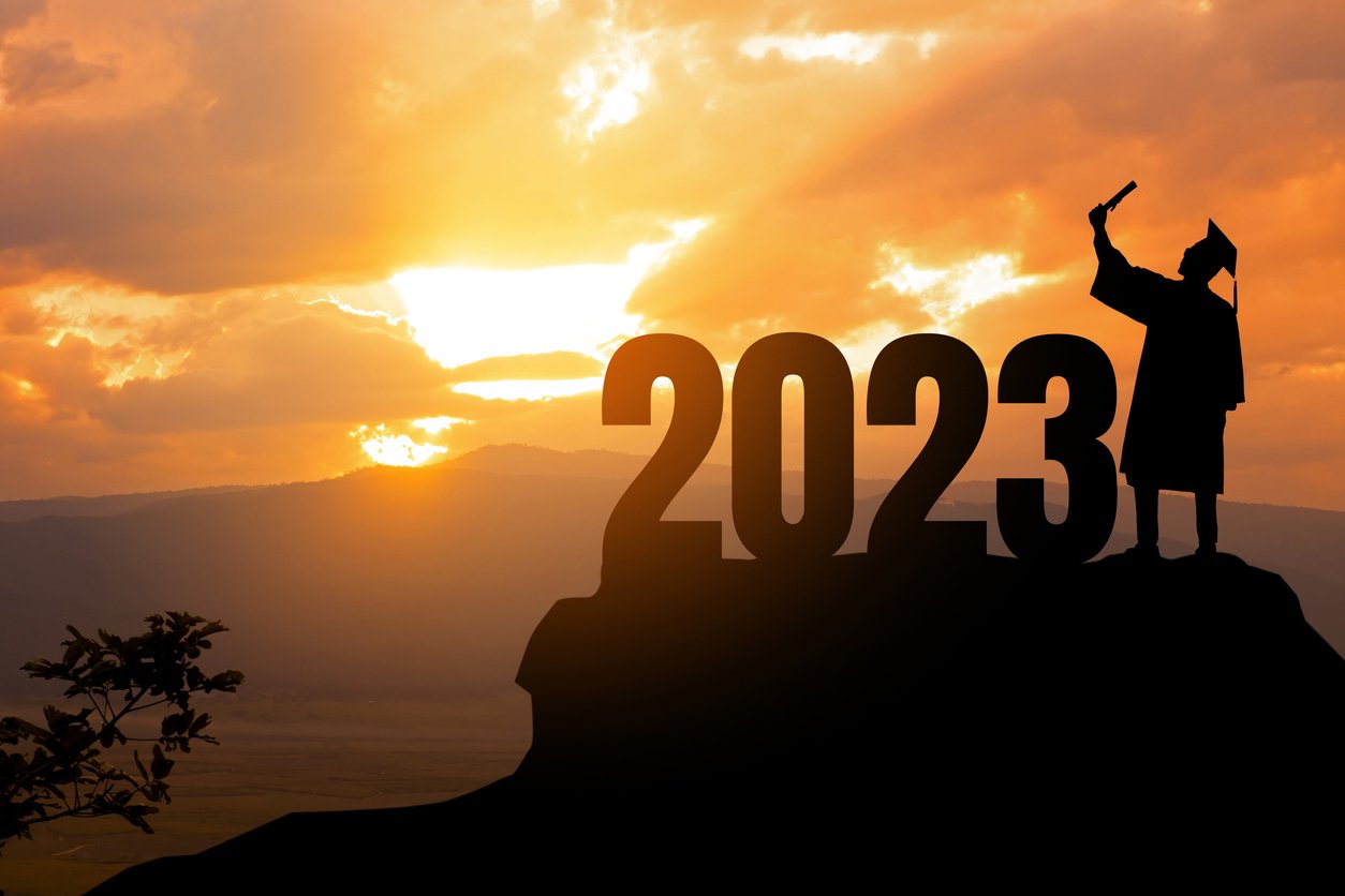 image shows a student standing beside a sign saying 2023 in his graduation cap and gowns with a sunset in the backrgound