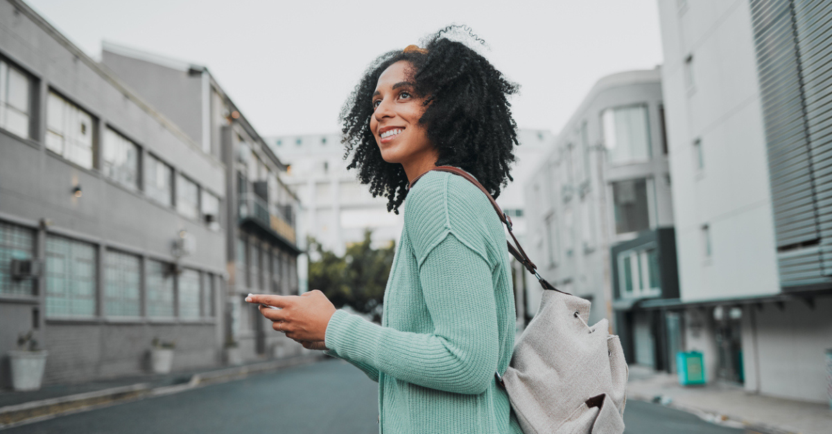 Female of color smiling with paper in hand in front of buildings