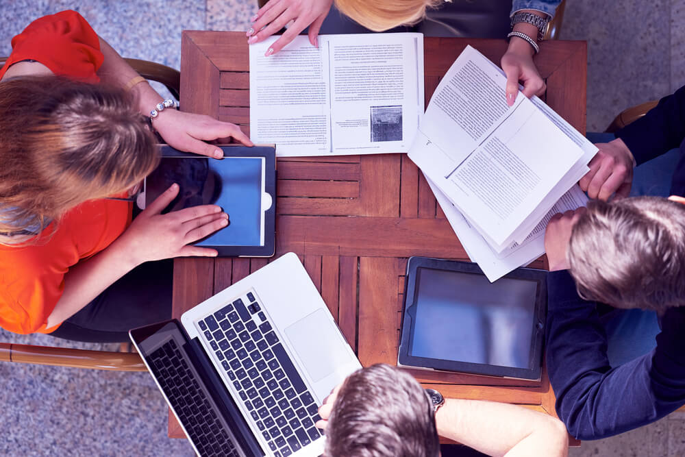 images shows students sitting around a desk with textbooks and laptops