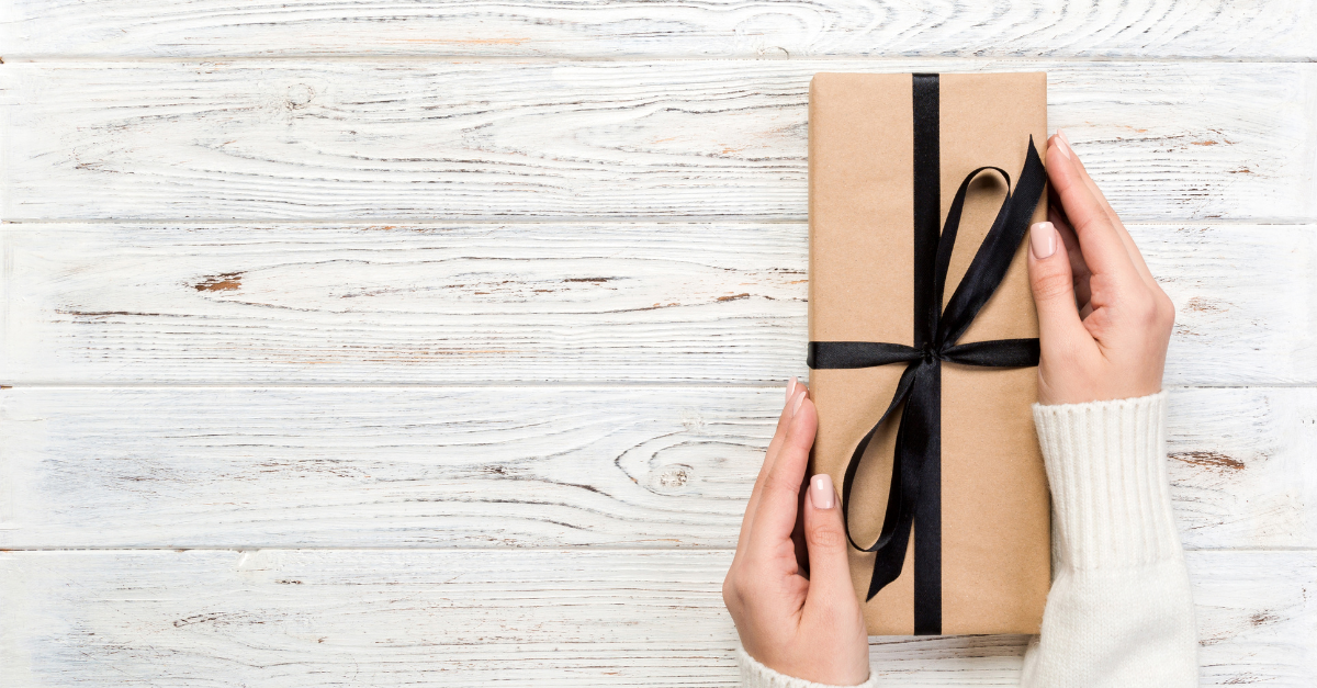 women's hands holding a brown paper covered box wrapped in a black ribbon against a background of whitewashed wood